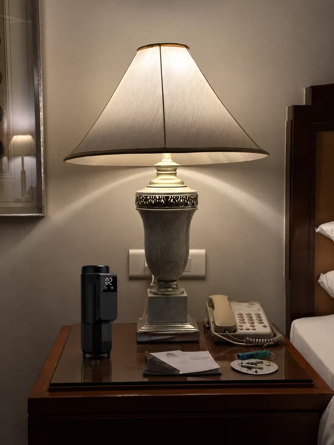 A classic table lamp with a beige fabric shade and a gray ceramic base sits on a dark wood bedside table in a hotel room setting, next to a phone, a water bottle, and a folded brochure.
